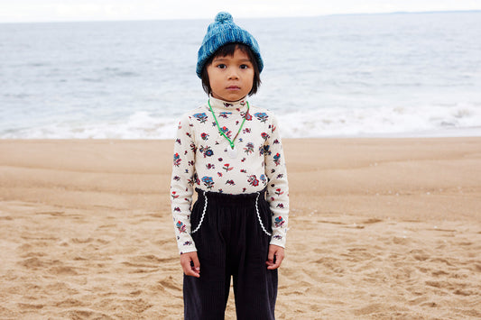 Child in colorful outfit and blue hat standing on beach.